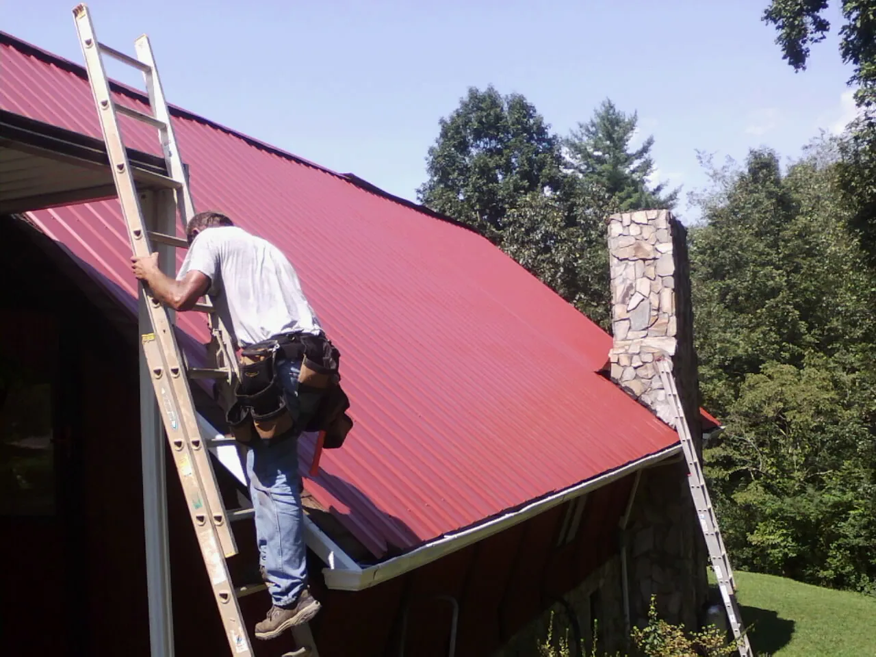 Roofer on a ladder beside a red metal roof and stone chimney — Soffit & Fascia Repair by Roofing Brothers in Platte City
